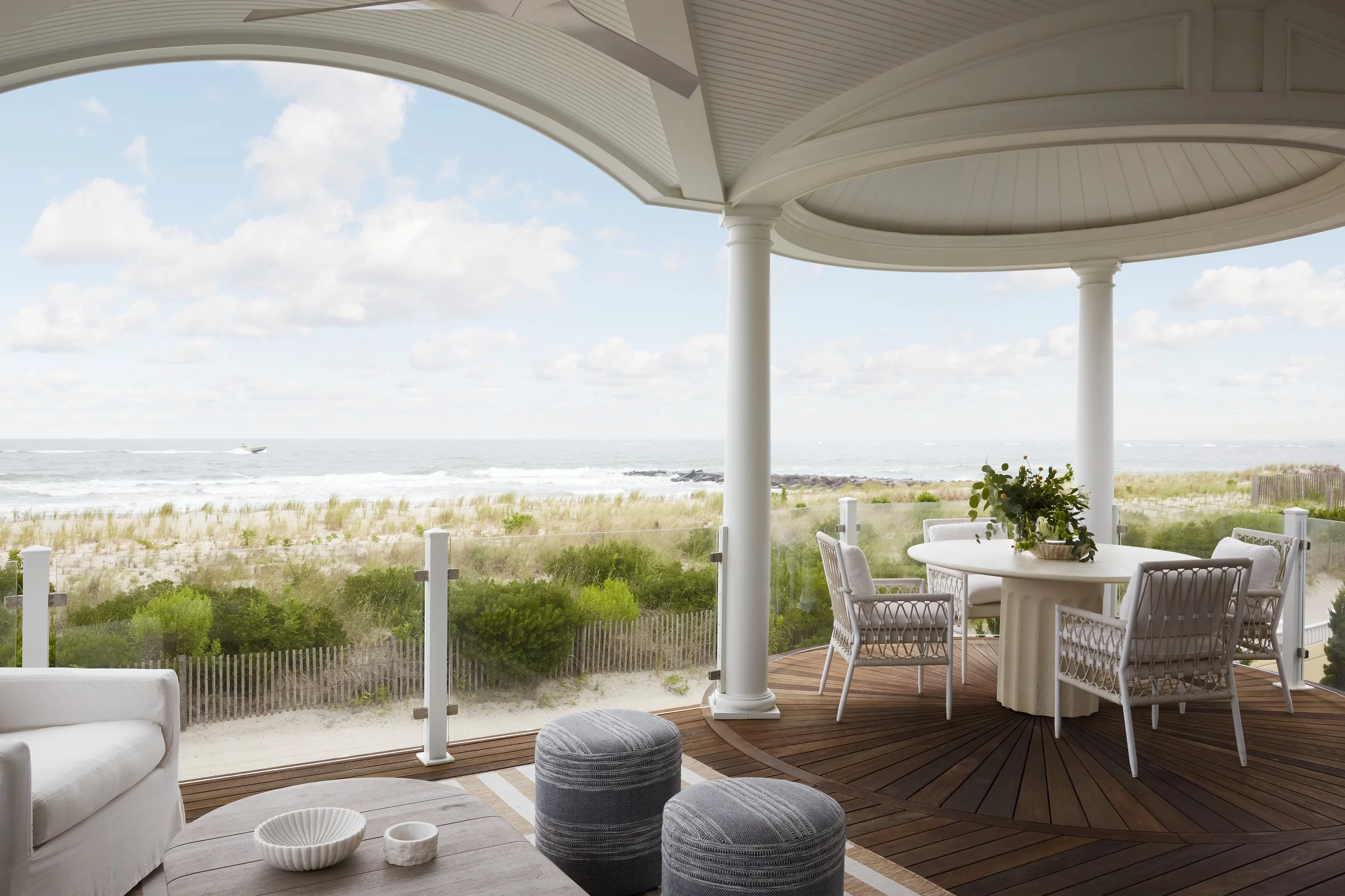 A seaside porch with white furniture, including a white sofa, a round table with a floral arrangement, and several chairs, overlooking a sandy beach and ocean.