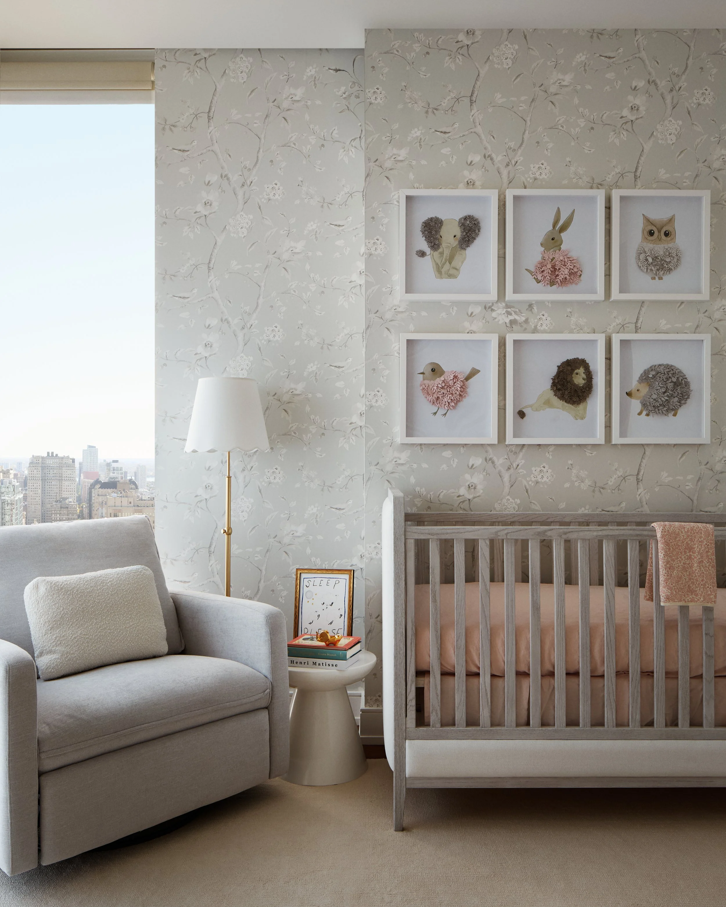 A cozy nursery room with a beige sofa, a small side table with books, a white floor lamp, a crib with a pink blanket, and framed artwork of animal illustrations on a floral wallpapered wall.