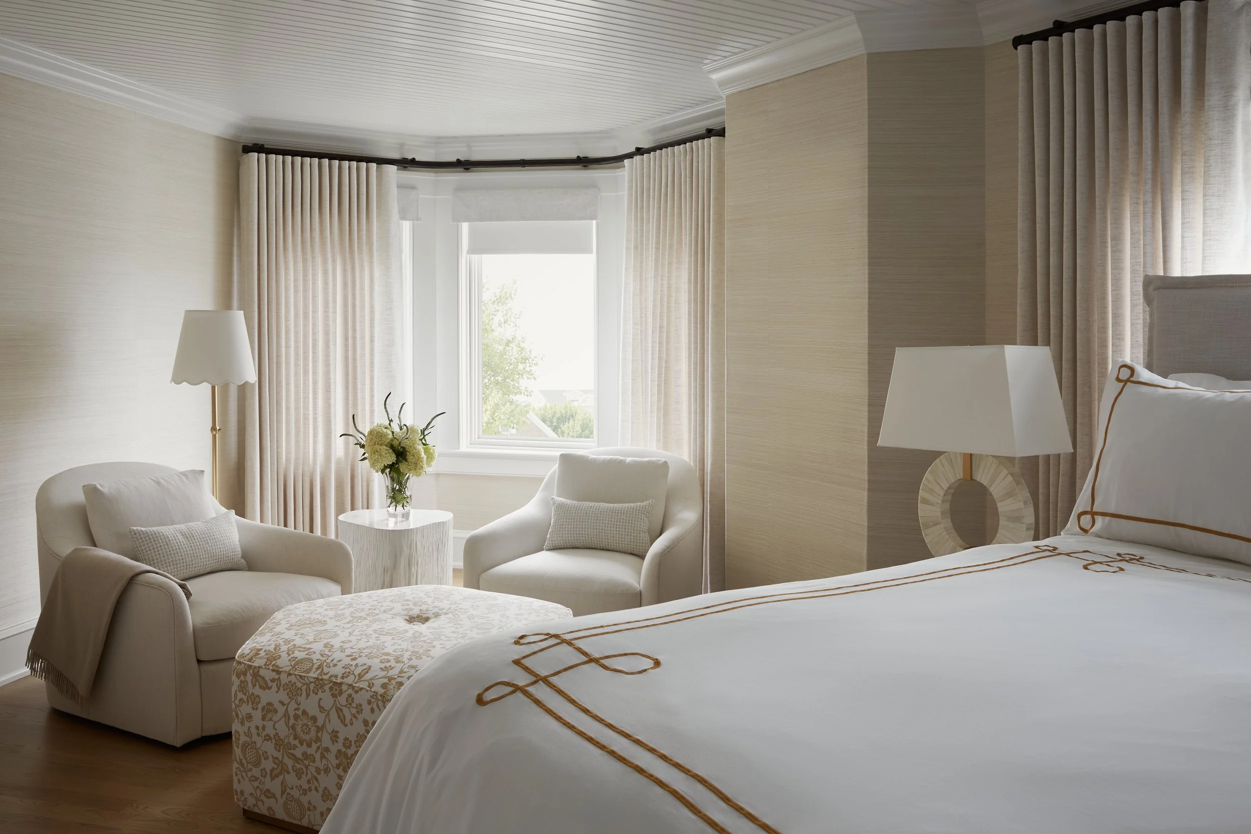 A bedroom with a large window, cream-colored curtains, a bed with white bedding, two cream-colored armchairs with pillows, a ottoman with a floral pattern, and a small table with a flower arrangement.