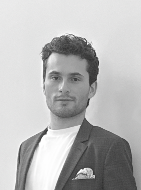 Black and white photo of a man with curly hair wearing a blazer and a white shirt, standing against a plain wall.