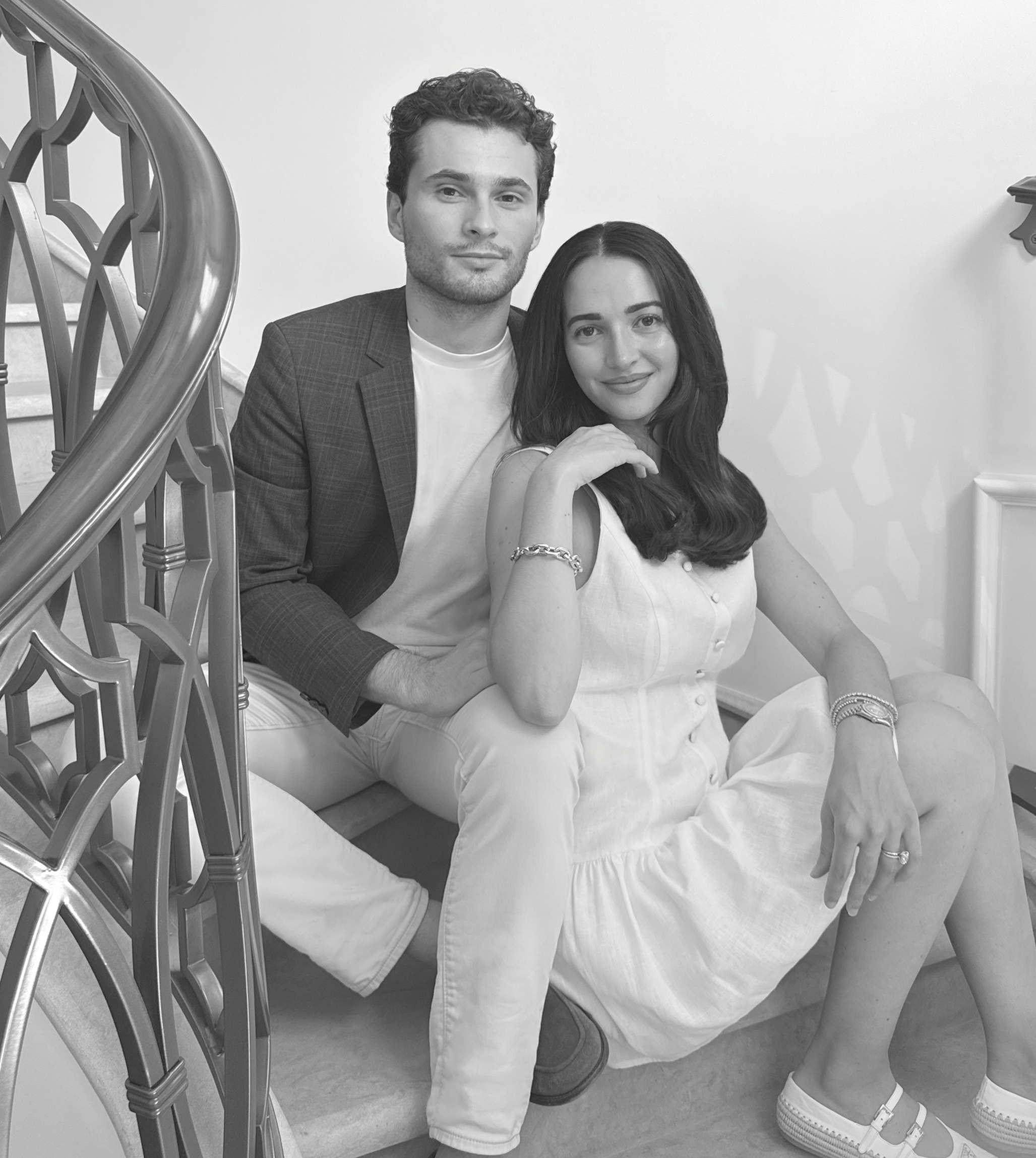A black and white photo of a young man and woman sitting on a staircase. The man is wearing a blazer and white shirt, and the woman is wearing a white dress with jewelry. They are posing for the camera with relaxed expressions.