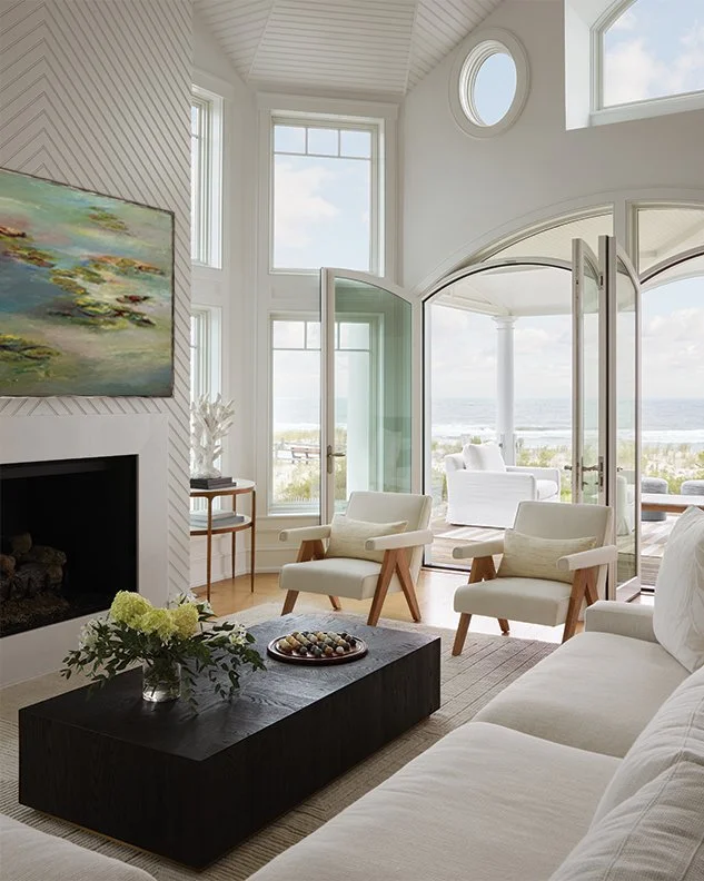 Living room with white walls and large windows, overlooking the ocean, featuring white armchairs, a gray sofa, a black coffee table with a flower vase, and a fireplace with a painting above it.