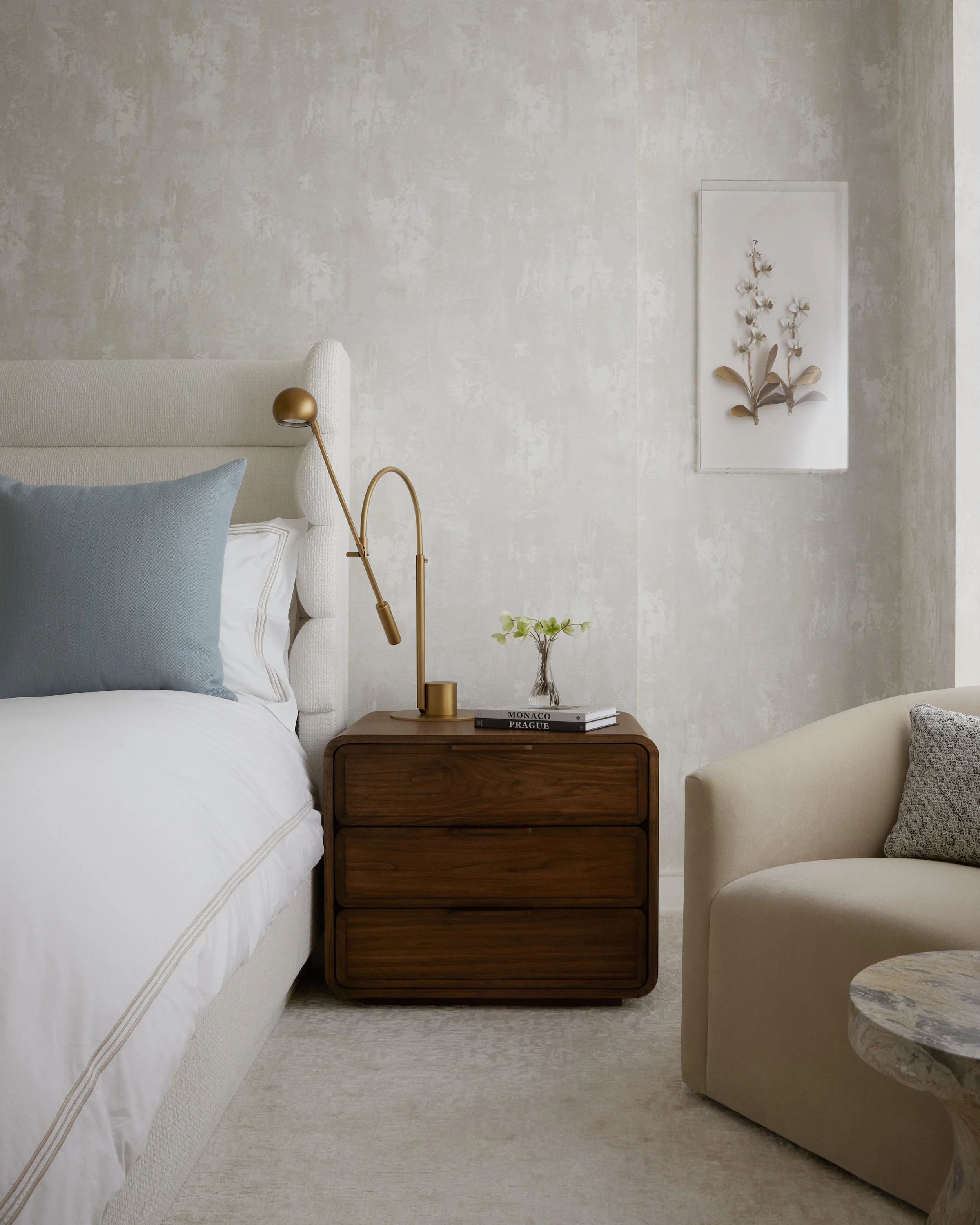 A bedroom corner with a beige upholstered bed and light blue pillow, a wooden nightstand with a modern gold lamp, a small vase with green leaves, and a stack of books, and a beige armchair with a patterned cushion, with a white textured wall and floral framed artwork in the background.