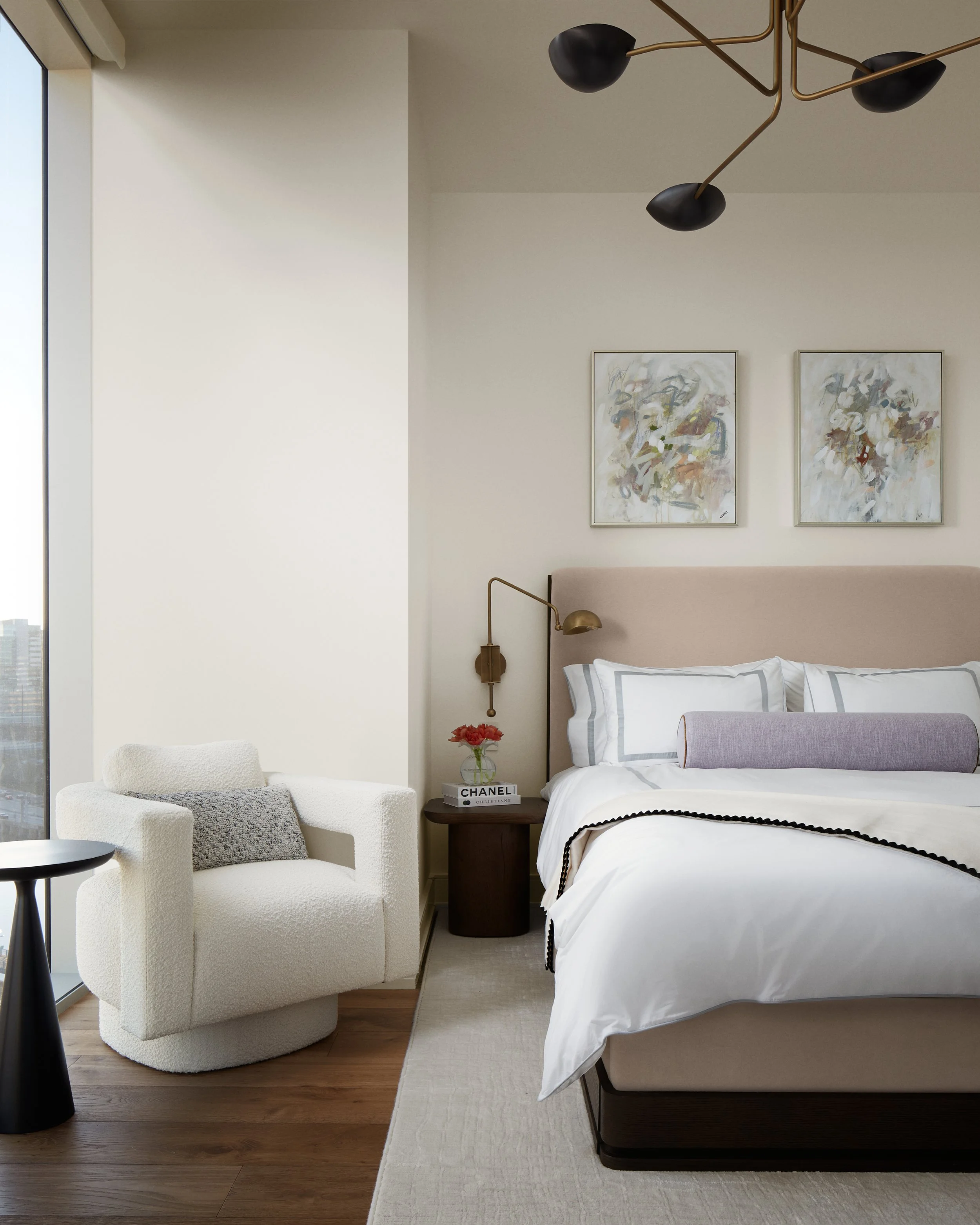 Modern bedroom with a large bed, abstract artwork on the wall, a bedside table with a vase of red flowers and books, an armchair with a pillow, a nightstand, and a modern black and gold ceiling light fixture.
