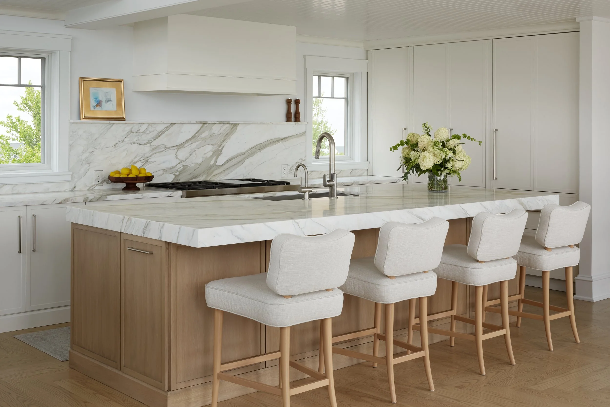 Modern kitchen with white marble countertops, island with four beige upholstered bar stools, white cabinetry, large flower vase, and two small windows showing greenery outside.