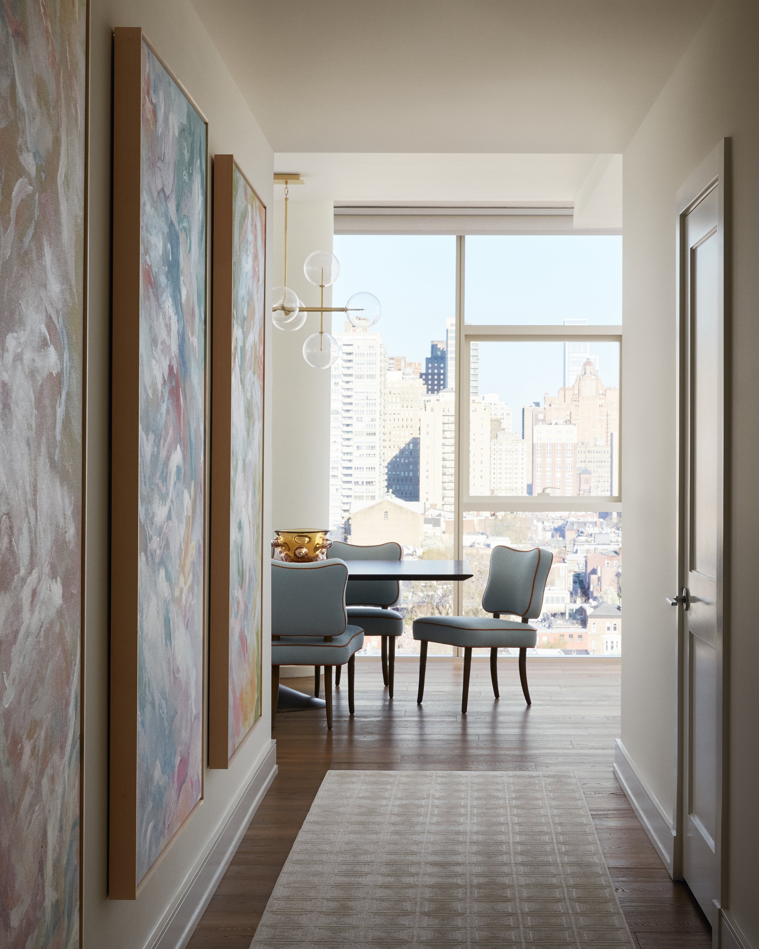 View of a dining area with a table and four light gray chairs with wooden legs, a large window showing a city skyline, hanging glass globe chandelier, and abstract paintings on the left wall.