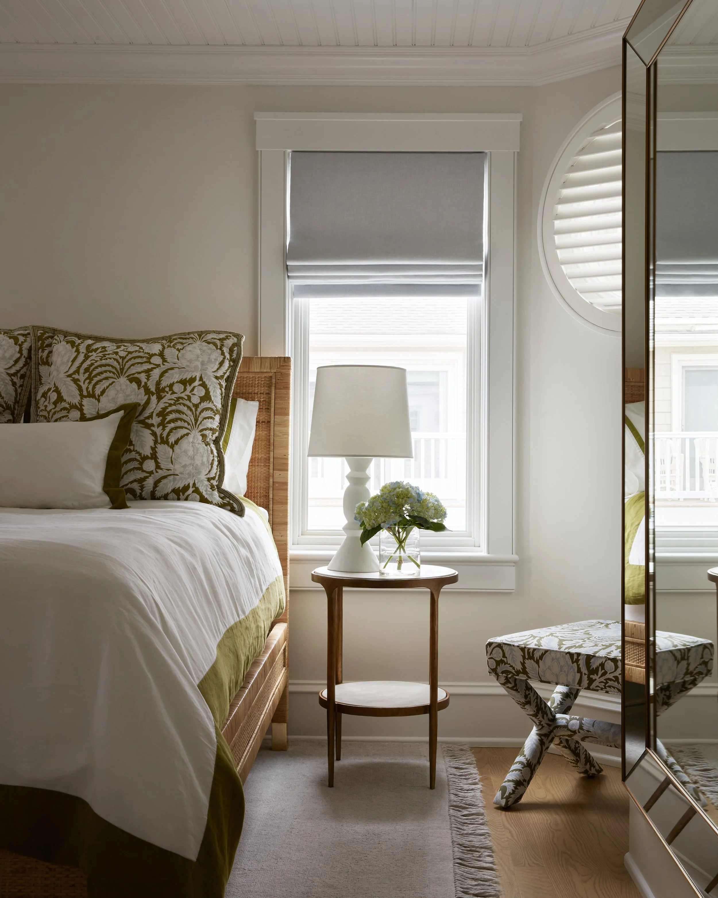 A cozy bedroom with a bed, bedside table with lamp and flowers, window with blinds, and a decorative stool.