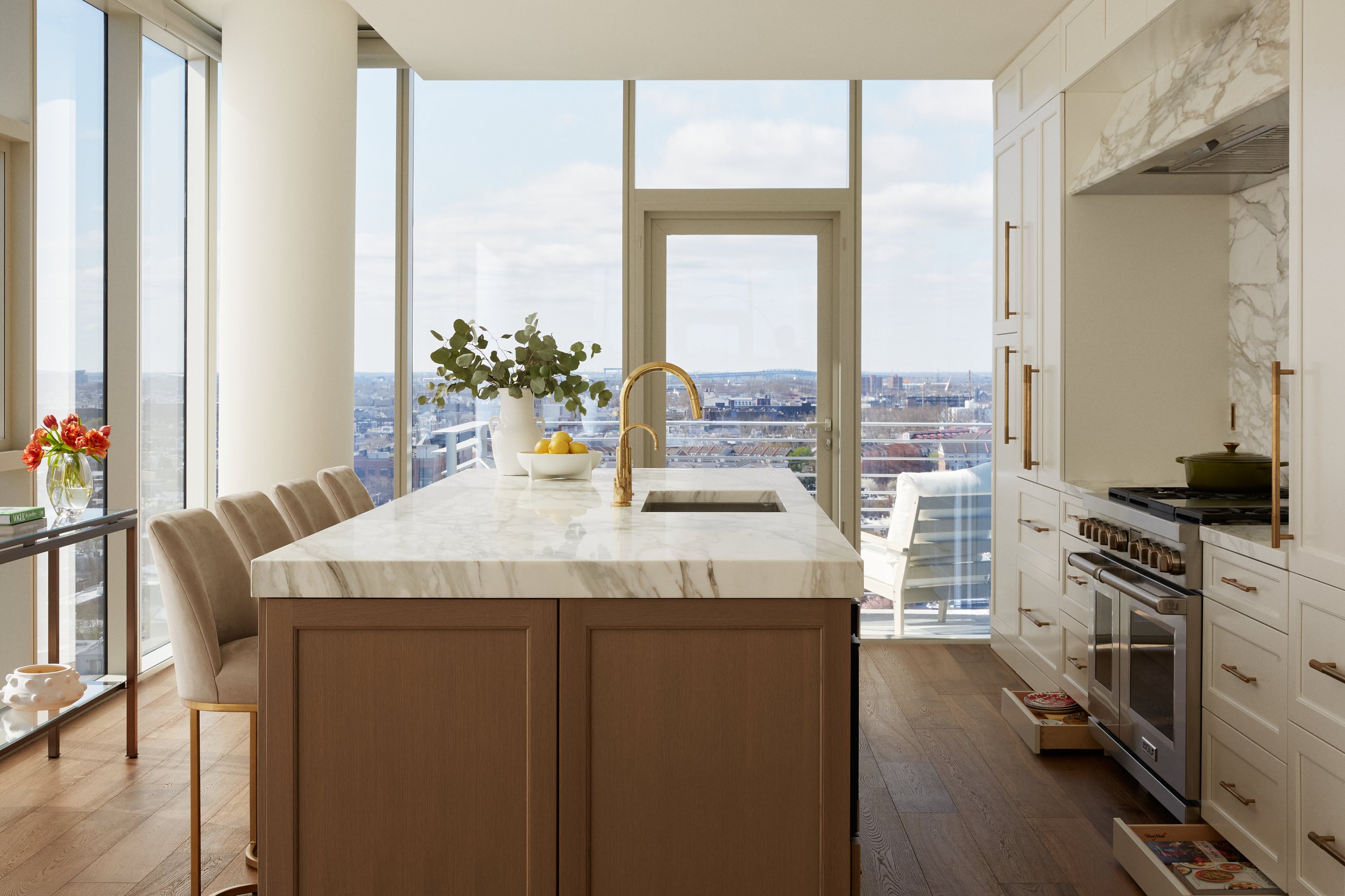 Modern kitchen with a central marble-topped island, beige chairs, large windows, and a view of the city skyline.