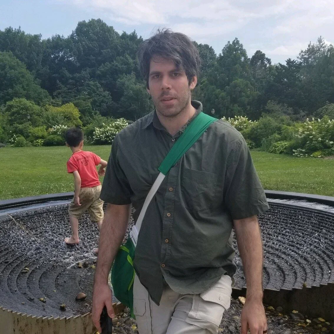 Man with dark hair and beard standing outdoors near a circular water fountain, with a young boy playing in the background, green trees, and cloudy sky.