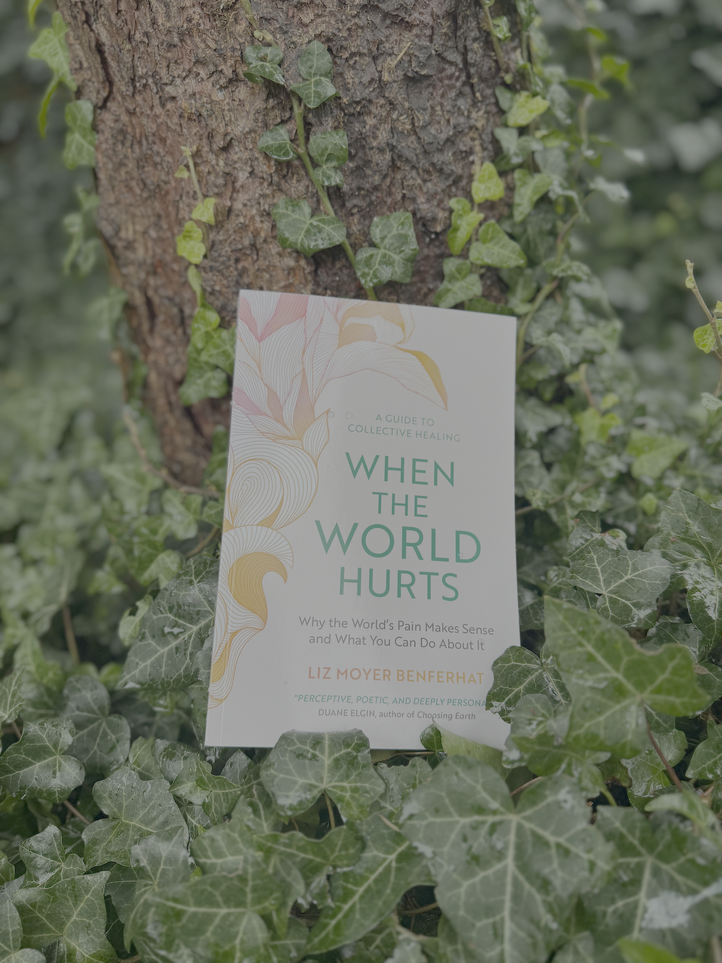 A book titled 'When the World Hurts' by Liz Moyer Benferhat leans against a tree trunk surrounded by green ivy.