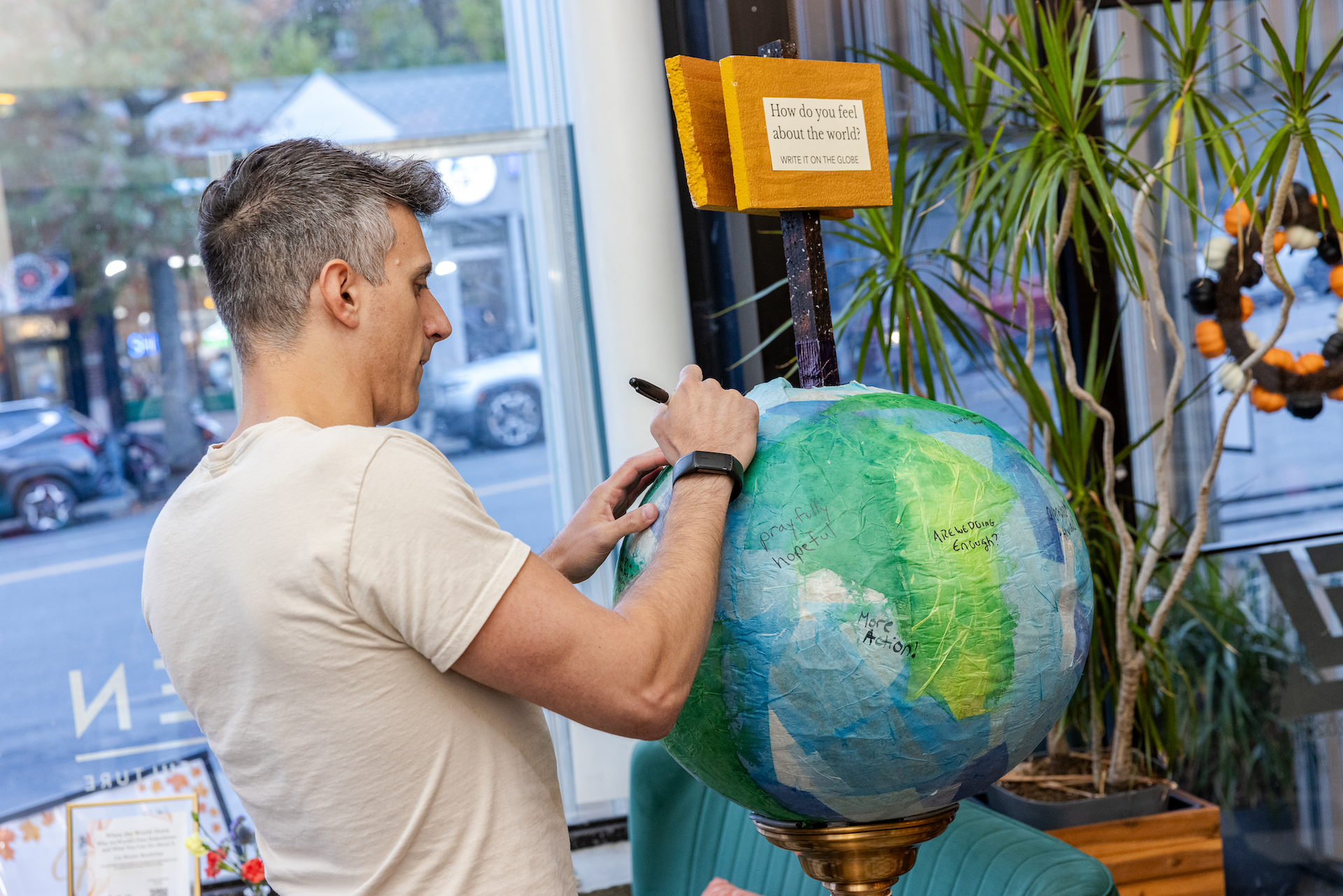 A man writing on a paper-mache globe of the Earth in a cafe, with a sign asking 'How do you feel about the world? WRITE IT ON THE GLOBE', surrounded by plants and large windows.