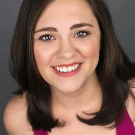 Close-up of a young woman with shoulder-length dark brown hair, smiling, wearing a pink top against a gray background.
