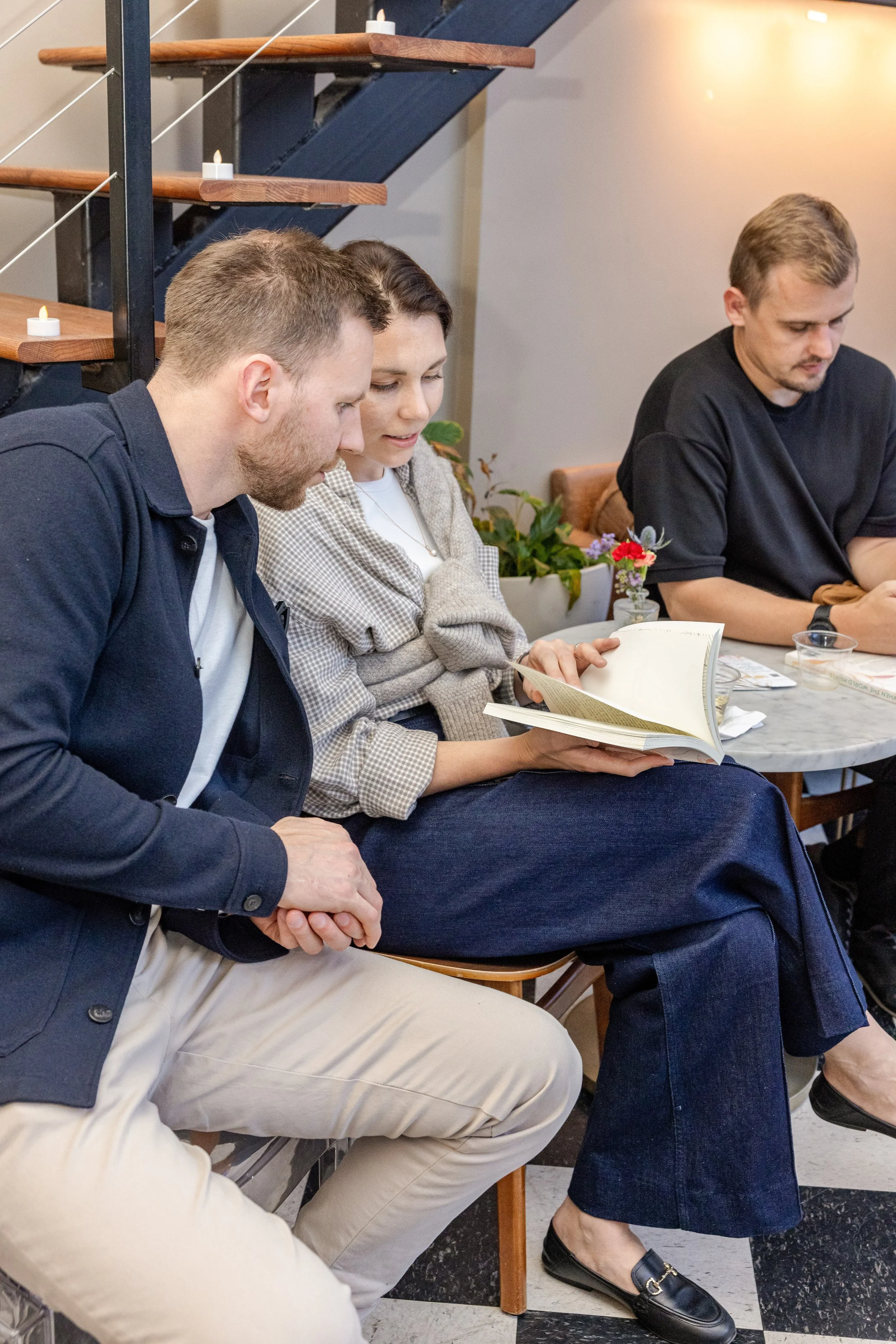 Three people sitting at a table, with a woman reading a book. The setting appears to be indoors with staircase decor and small candles on the staircase to the left.