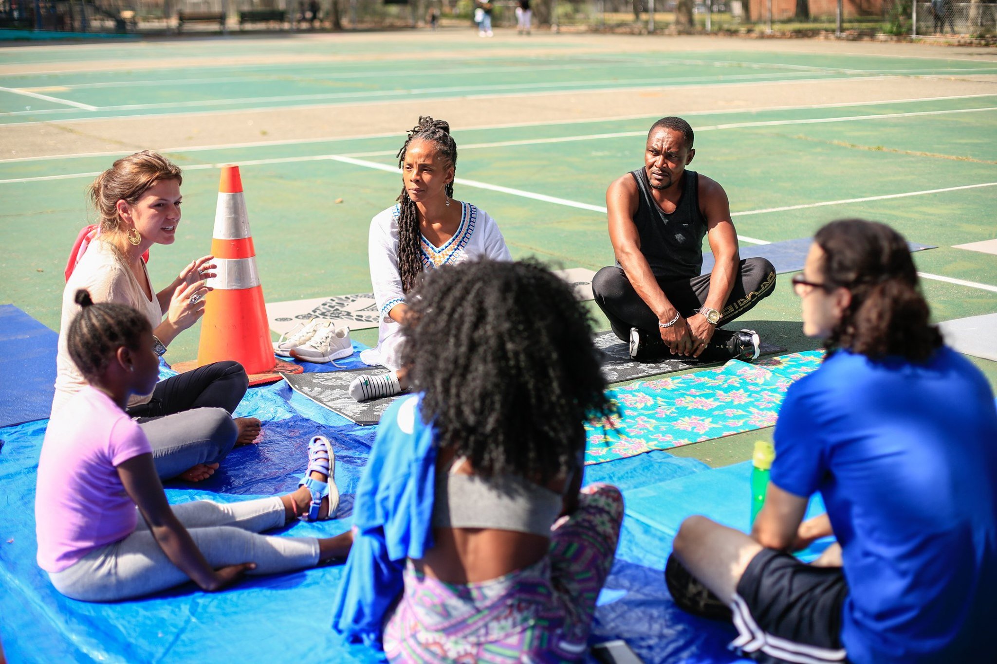 A group of people sitting on mats on an outdoor tennis court, engaged in a discussion or workshop, with an orange traffic cone near them.