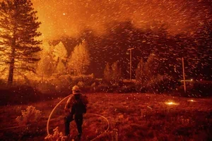 A firefighter spraying water on a wildfire at night with flames and smoke in the background.