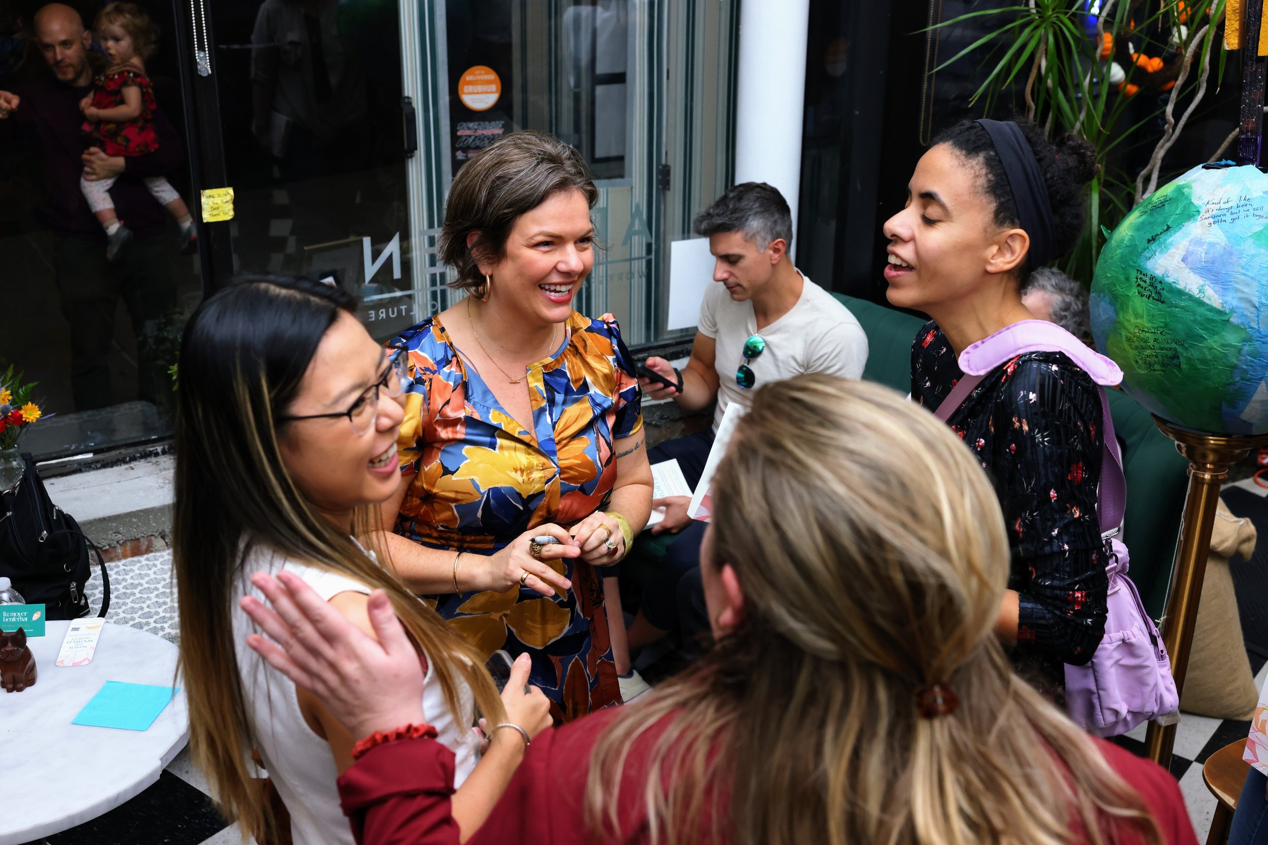 Group of smiling women and a man engaging in conversation at a social gathering in a cozy indoor space. One woman is wearing glasses, another has a backpack, and a man is looking at his phone. A globe is visible in the background.