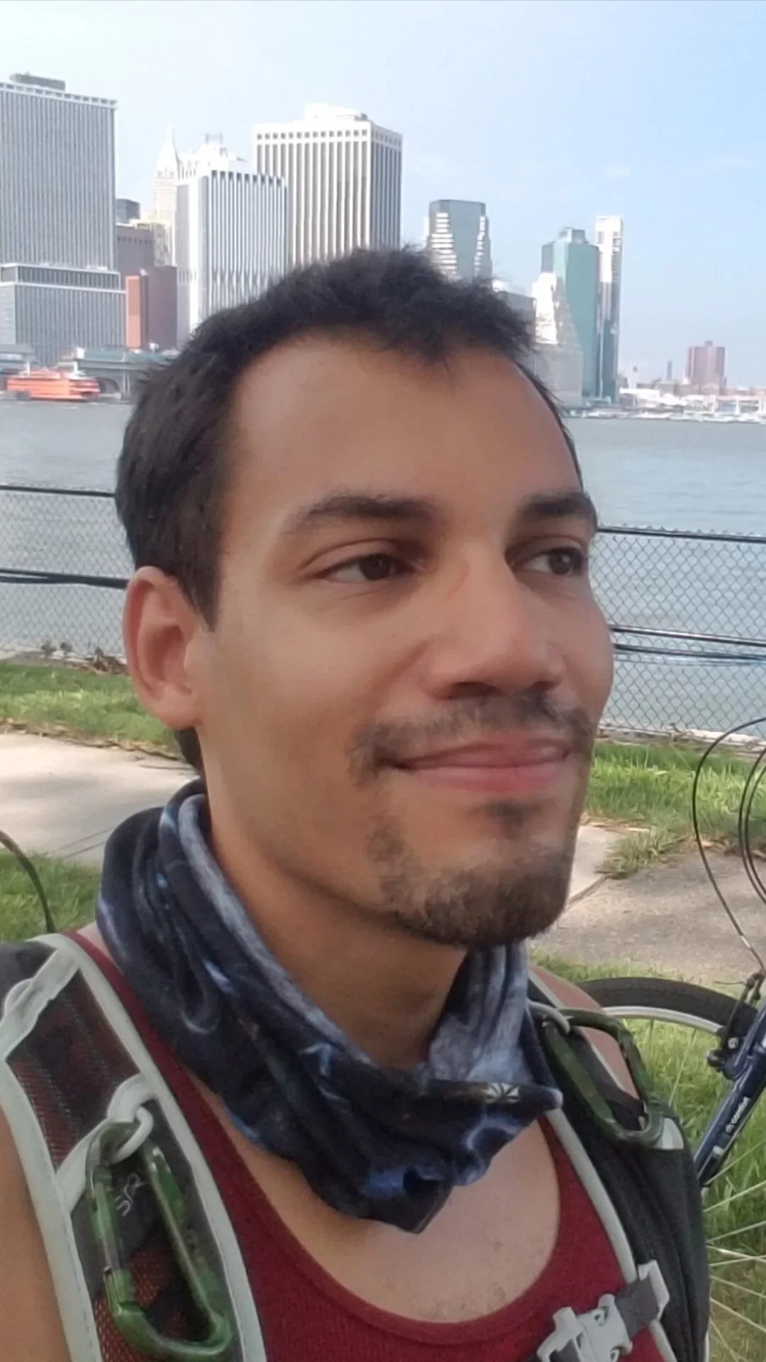 Close-up of a man with short dark hair, a goatee, and a marathon running vest, smiling outdoors with city skyscrapers and water in the background.