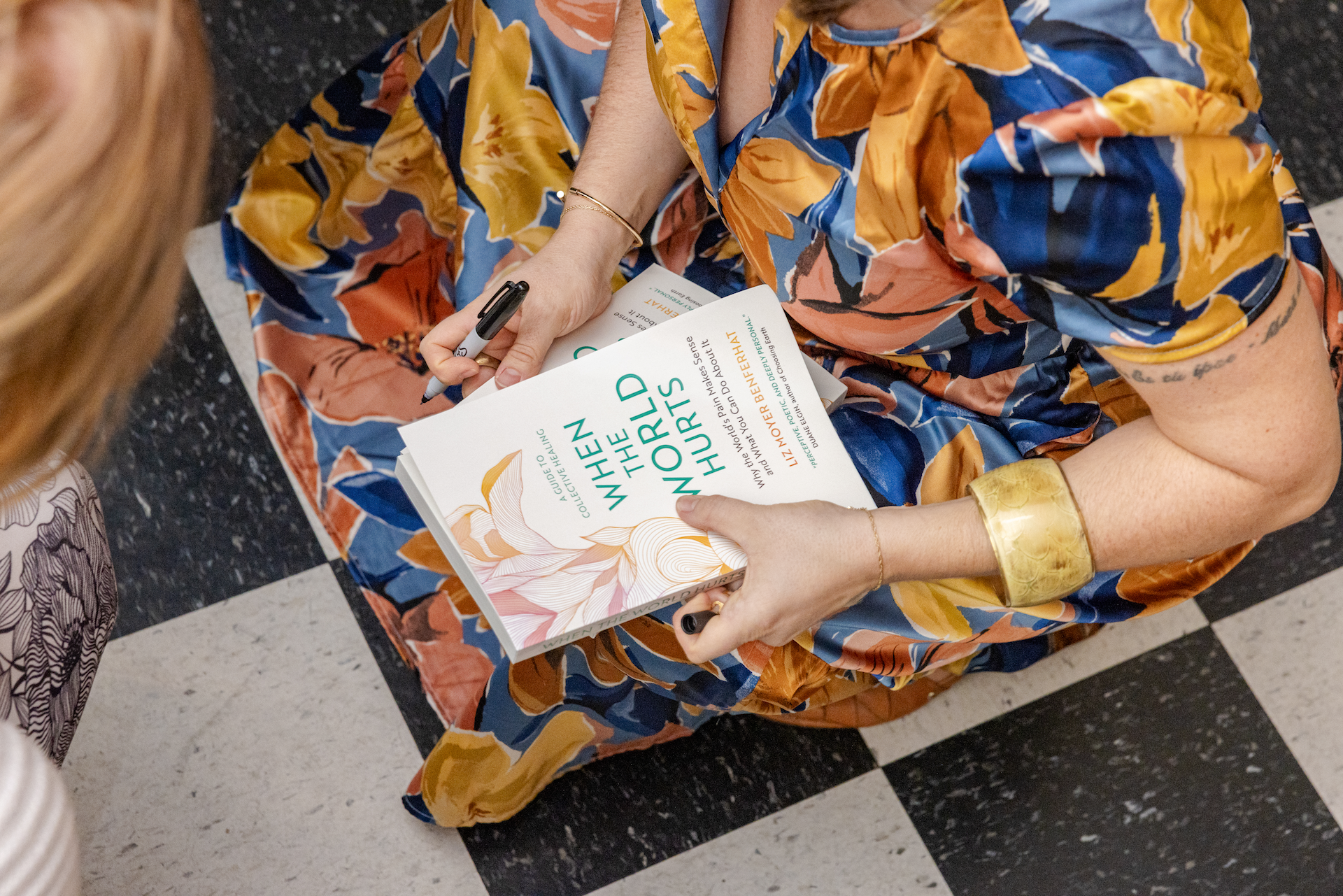 A woman in a colorful floral dress is sitting on a black-and-white checkered floor, holding a book titled 'When the World Hurts' by Liz Moore. She has a black marker in her right hand and wears a gold bracelet and a delicate gold bracelet on her left wrist.