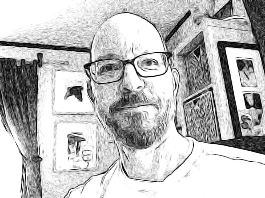 A black-and-white stylized portrait of a bald man with glasses and a beard, standing in front of a wall with framed pictures and artwork.