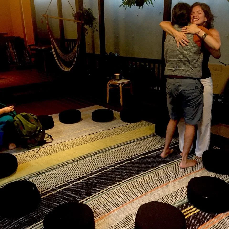 Two women hugging and smiling in an indoor setting with a hammock and some seating options in the background.