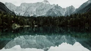 A calm mountain lake with trees along the shoreline and snow-capped mountains in the background, reflected in the water.