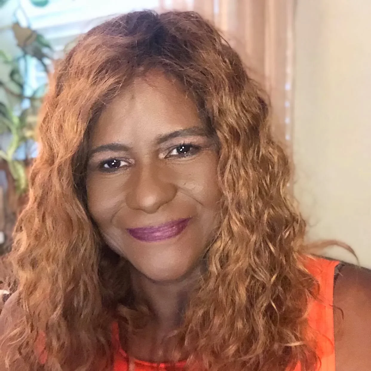 A woman with long, curly, reddish-brown hair smiling in an indoor setting.