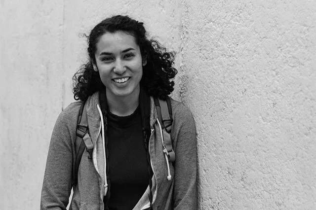 A young woman with curly hair smiling, wearing a backpack and casual clothes, standing against a textured wall.
