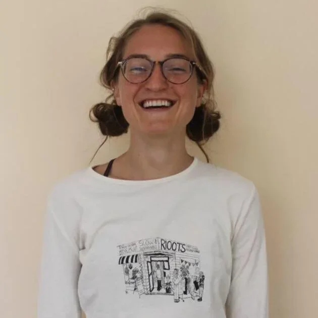 A woman smiling with glasses, wearing a white T-shirt with a black and white illustration of a storefront with the word 'ROOTS' on it.