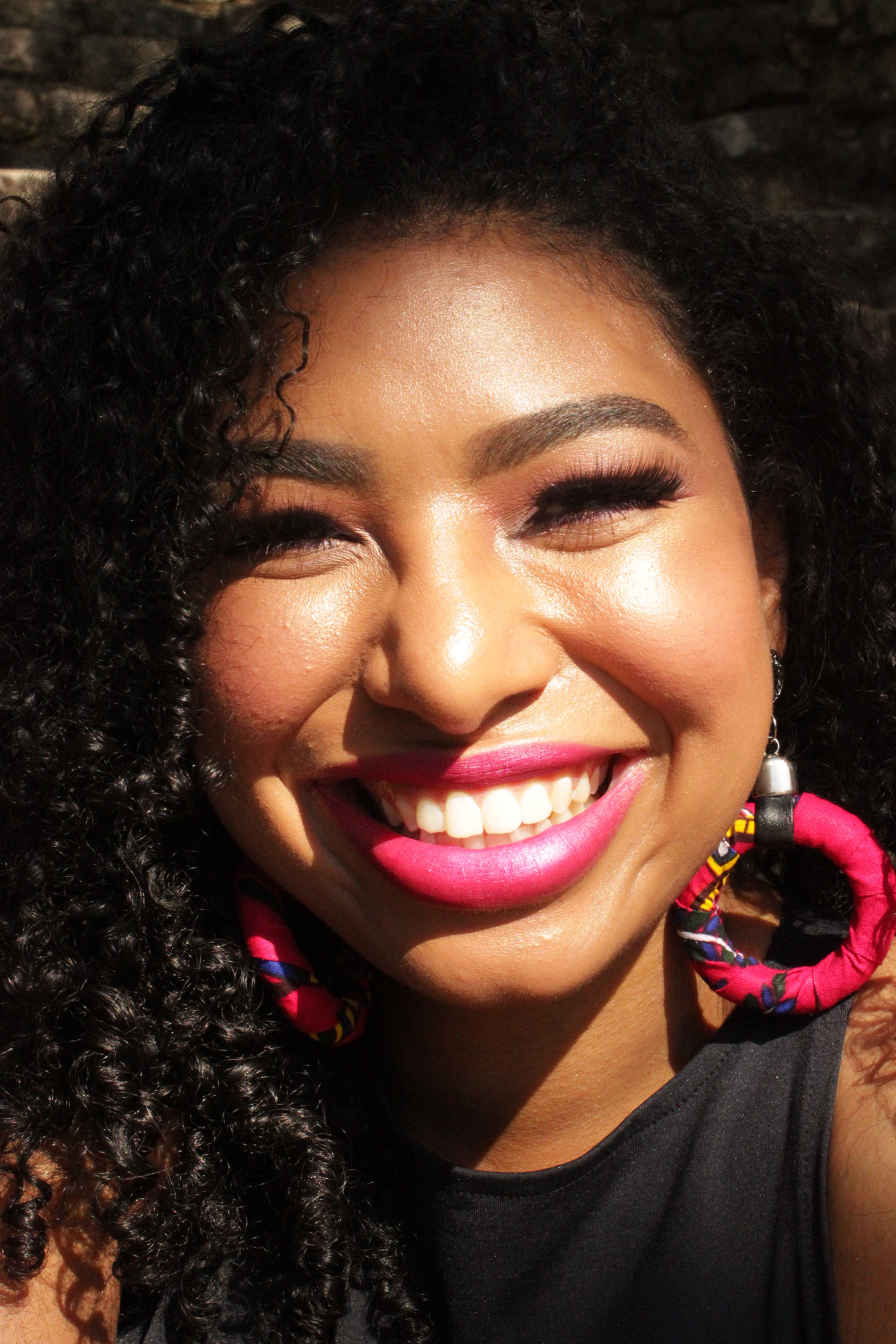 Close-up of a smiling woman with curly hair, pink lipstick, and colorful earrings.