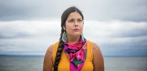 A woman with a braid wearing a yellow top and a pink and purple bandana, outdoors with a cloudy sky and water in the background.