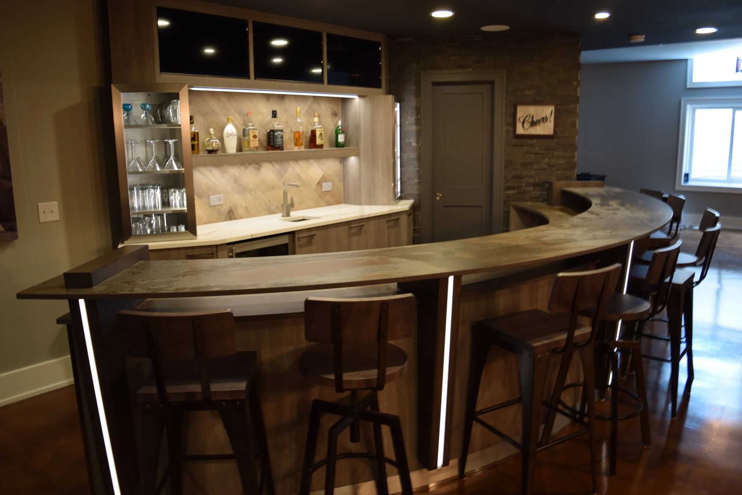 Wooden bar with barstools, liquor bottles, glassware, and a sink, in a dimly lit room.