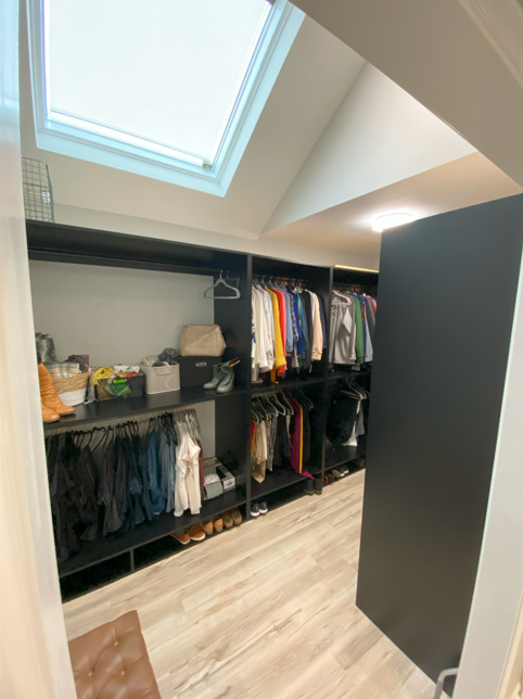 Walk-in closet with black shelves filled with clothing, shoes, and storage boxes, under a skylight