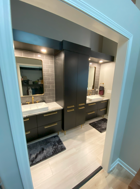 Modern bathroom with black cabinets, gold handles, two white vessel sinks, large mirrors, gray tiled wall, and two black rugs on a light-colored wood floor.