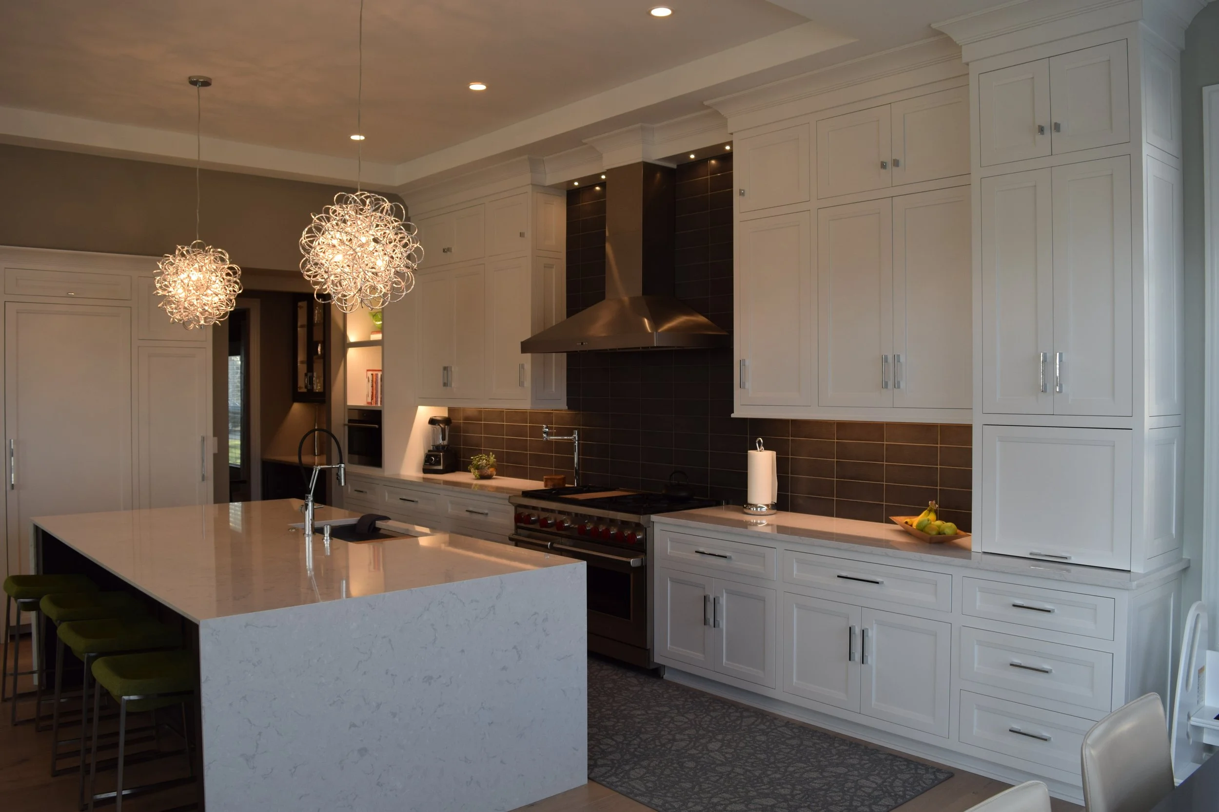 Modern kitchen with white cabinets, dark tile backsplash, stainless steel range hood, island with sink, and unique pendant lighting.