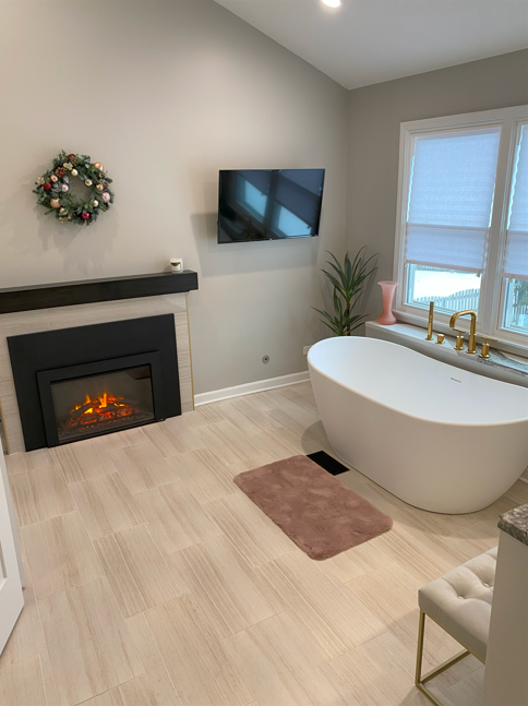 A modern bathroom with a freestanding bathtub, a fireplace, a wall-mounted TV, a decorative wreath, a potted plant, a window with blinds, a pink vase, and a pink bath mat.