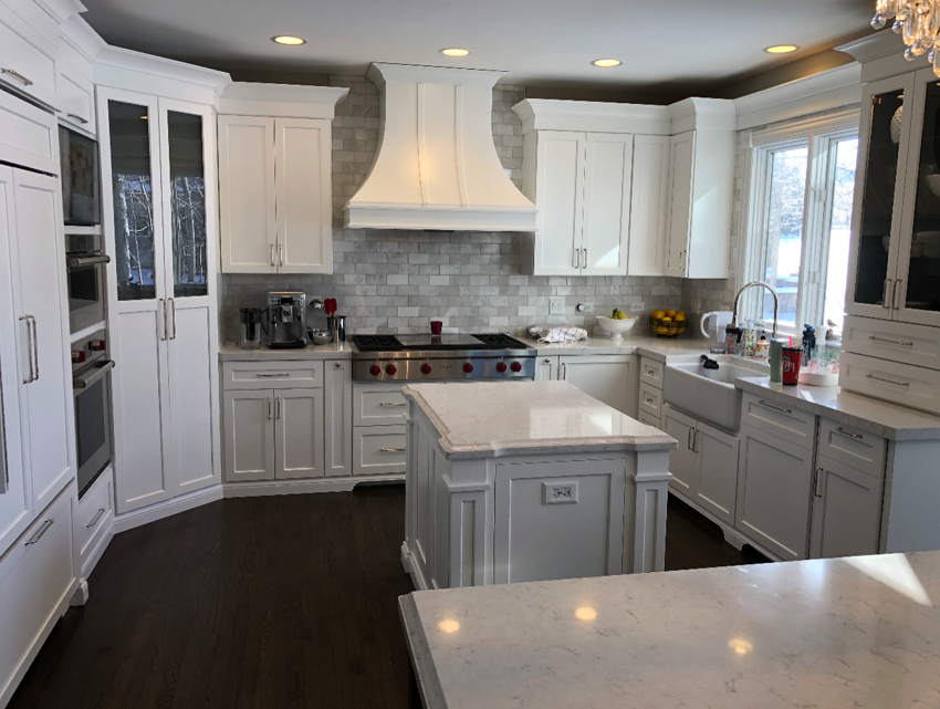Modern white kitchen with island, brick backsplash, stainless steel appliances, and a sink under a window.