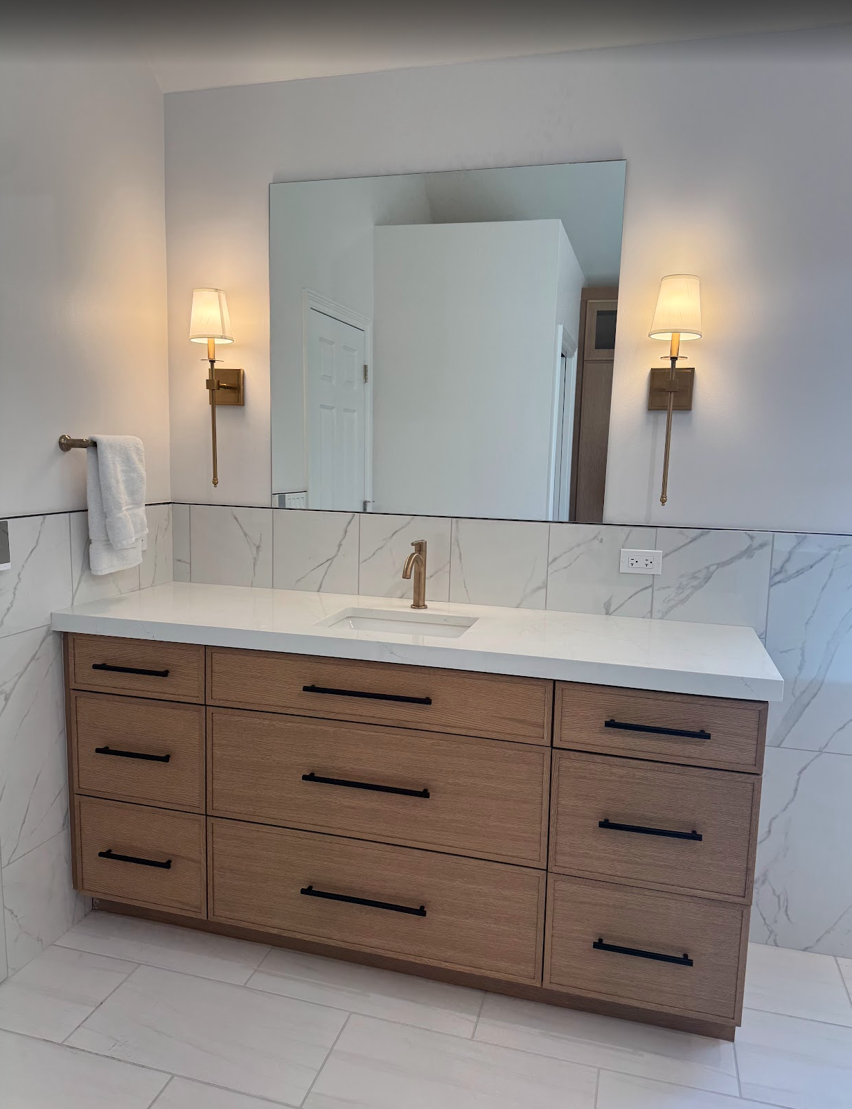 Modern bathroom vanity with a wood cabinet, white marble countertop, and a gold faucet, reflected in a large mirror flanked by wall sconces, with a towel on a towel bar and an electrical outlet.
