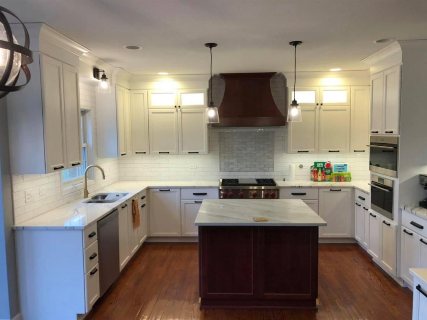 Modern kitchen with white cabinets, a central island with dark wood base, marble countertops, a dual oven, and a gas stove with a wooden range hood. Pendant lights hang above the island, and there is a backsplash with white subway tiles.