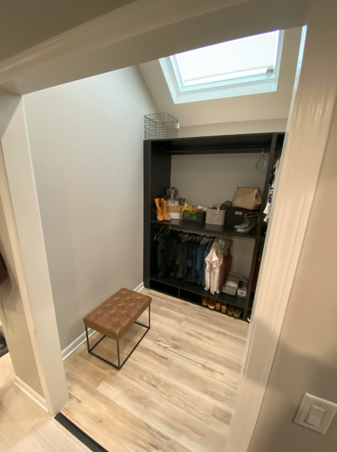 Small walk-in closet with black shelving unit, clothes hanging in the middle section, shoes at the bottom, and various storage boxes and accessories on top. A skylight window above provides natural light, and a small padded stool with metal legs is i