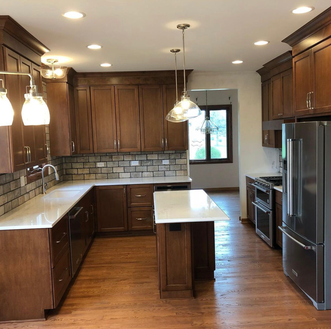 Modern kitchen with wooden cabinetry, white countertops, brick backsplash, stainless steel appliances, and pendant lighting over an island.