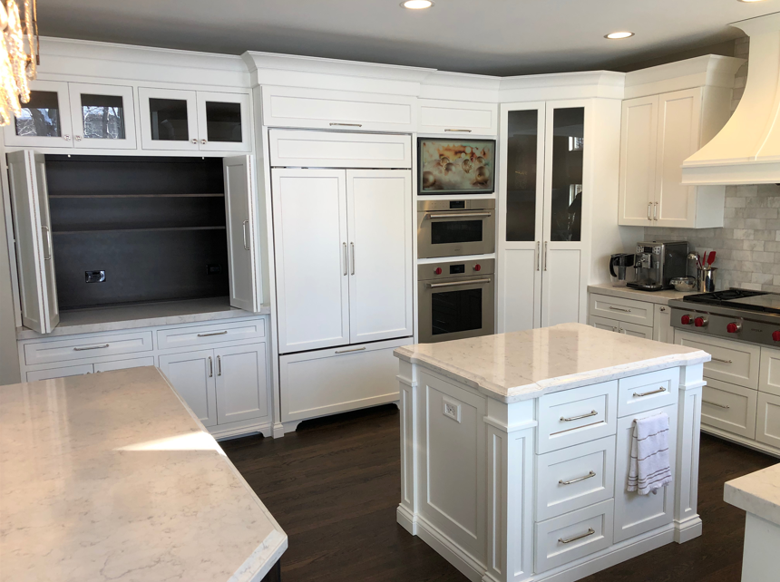 Modern kitchen with white cabinets, a corner island, marble countertops, built-in oven, microwave, refrigerator, and a white range hood.
