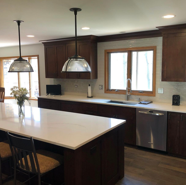 Modern kitchen with dark wooden cabinets, a large white island counter, pendant lights, a window above the sink, and stainless steel appliances.