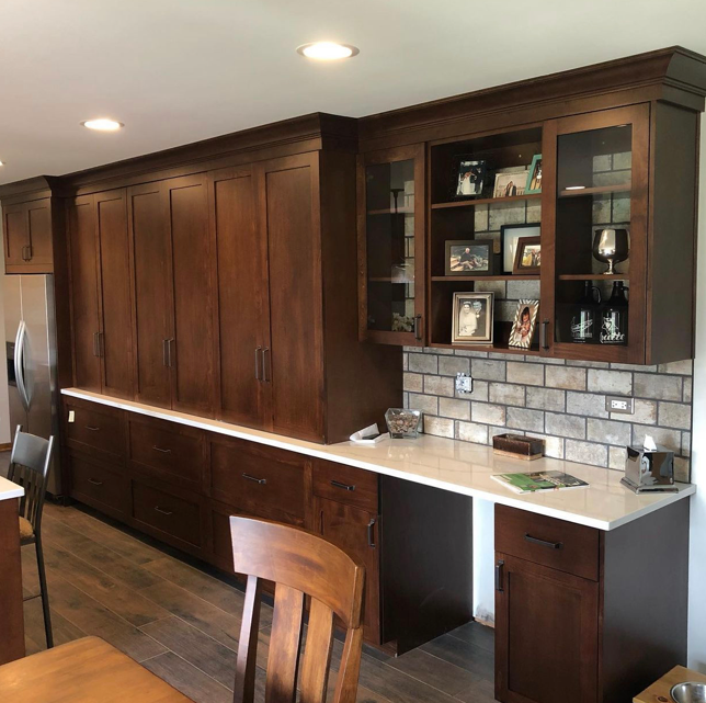 Kitchen with dark wood cabinetry, a light-colored countertop, and a brick backsplash. There are open shelves with framed photos and decorative items, and part of a dining table with wooden chairs is visible.