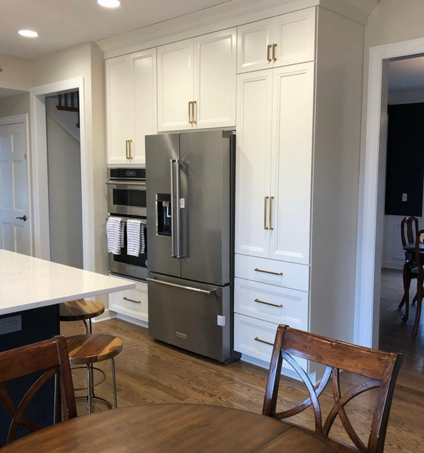 Kitchen with white cabinets, stainless steel refrigerator, built-in ovens, and a marble countertop island. Wooden chairs and a wooden stool are visible.
