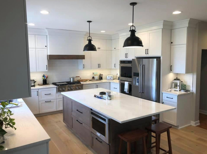 Modern kitchen with white cabinets, stainless steel refrigerator, black pendant lights, and a central island with a microwave. Wooden stools are positioned at the island, and various appliances and kitchen items are on the countertops.
