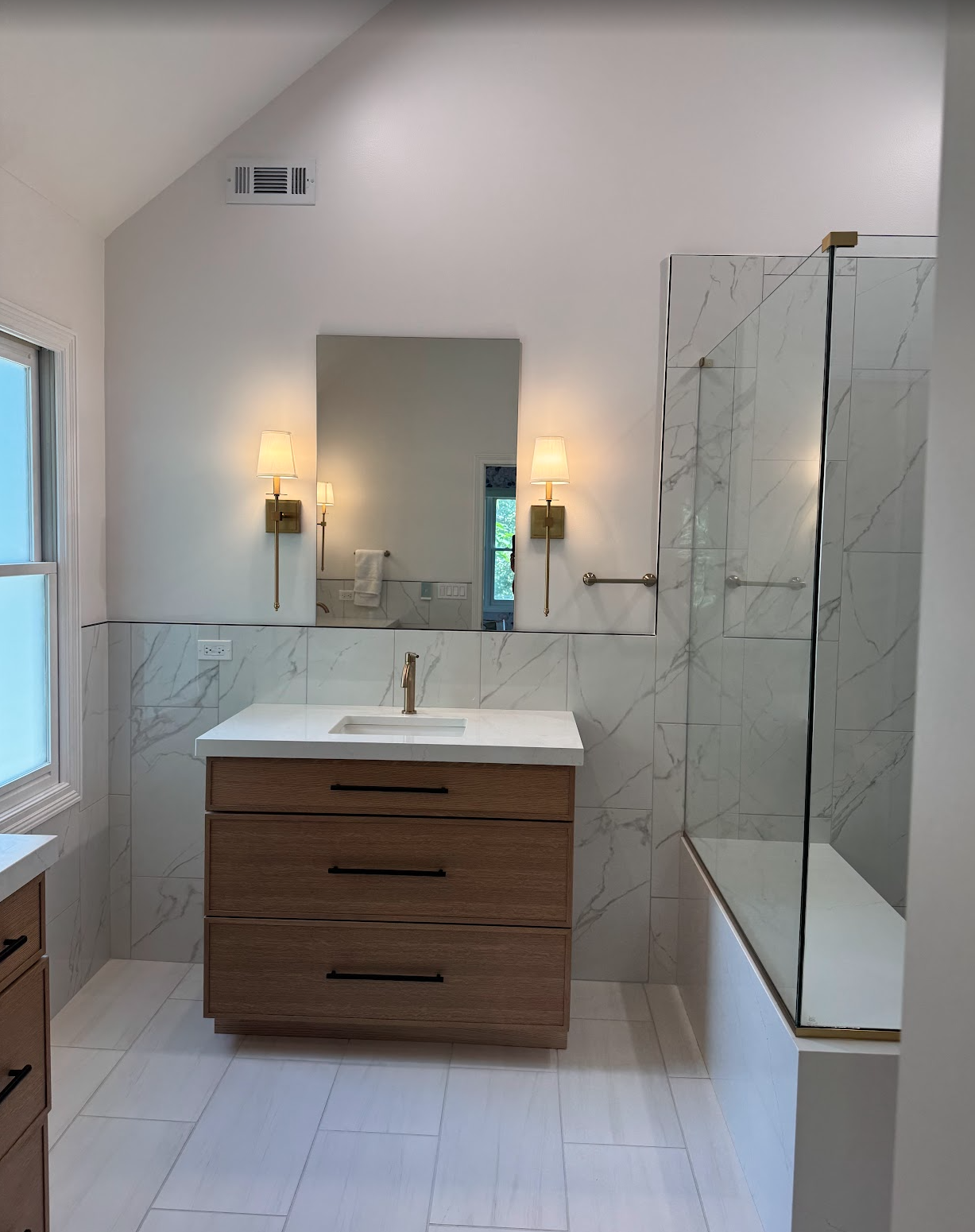 Modern bathroom with wooden vanity, white marble countertop, large mirror, wall-mounted lights, and glass-enclosed shower.