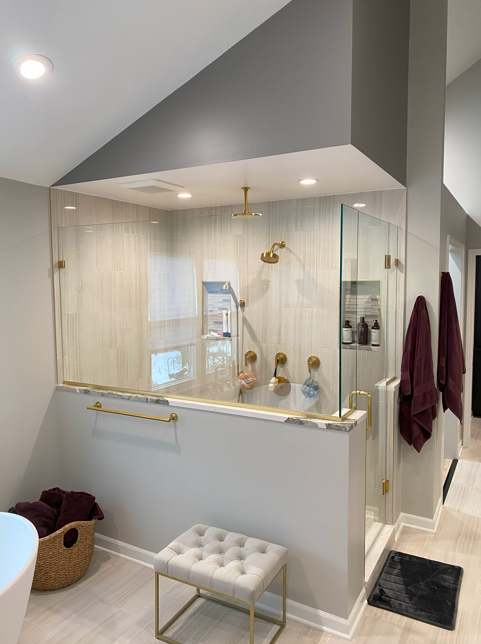 Modern bathroom with a walk-in shower surrounded by glass, featuring gold fixtures including a showerhead and handles. The shower has a built-in niche with toiletries. There are two maroon towels hanging on the wall, a small cushioned bench near the 