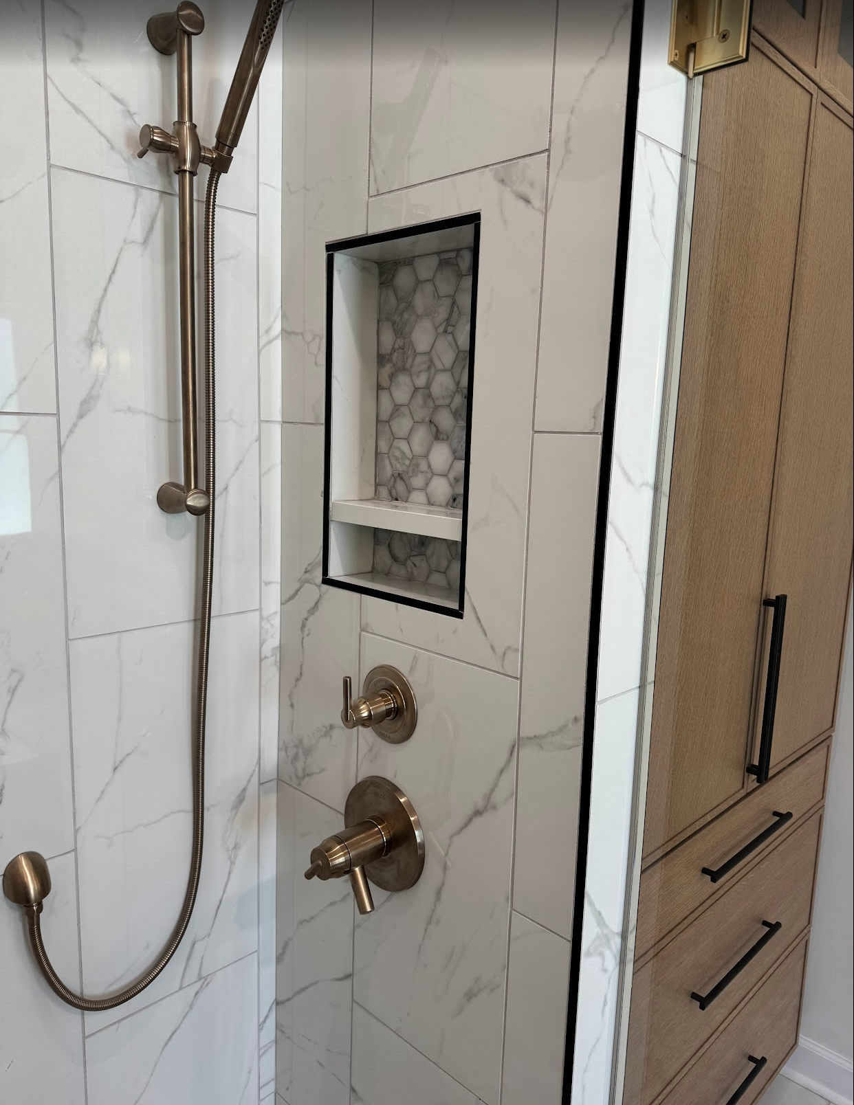 Modern shower with white marble tiles and a built-in niche with gray and white hexagonal tiles, along with bronze fixtures and a wooden cabinet nearby.
