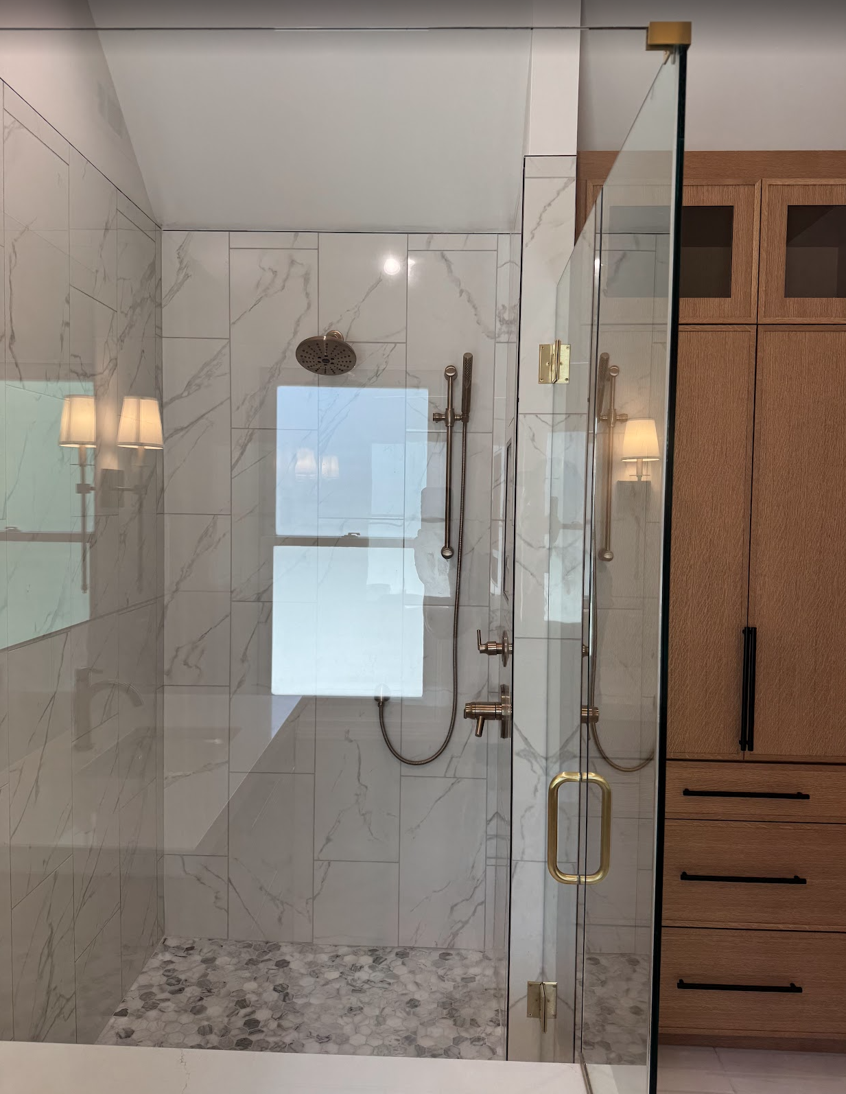 A modern glass-enclosed bathroom shower with white marble tiles, a rain shower head, a handheld shower, and a wooden cabinet beside it.