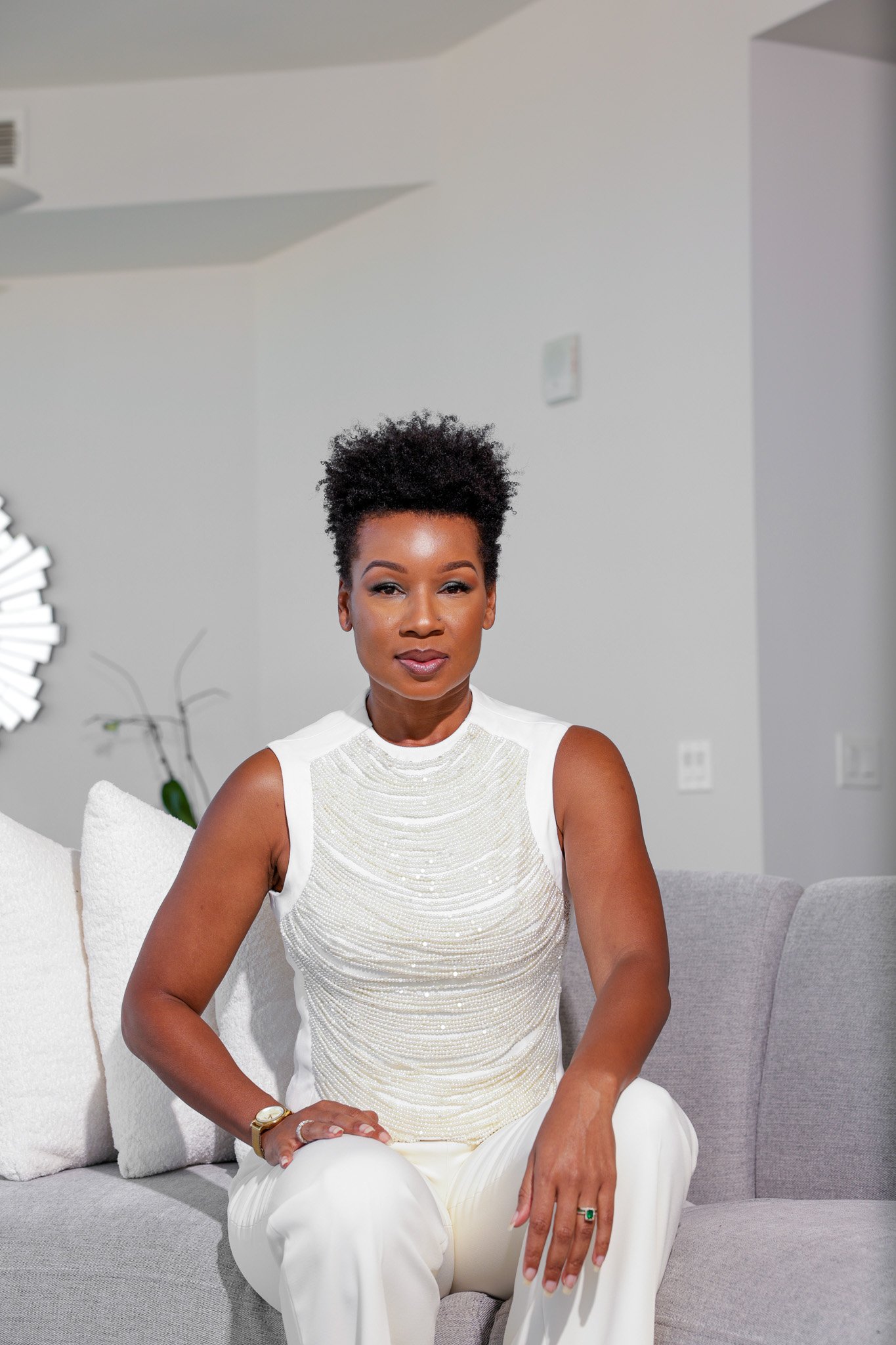A woman with short curly hair sitting on a light-colored sofa, wearing a white sleeveless top and white pants, with a watch on her left wrist and a ring on her right hand, in a modern living room.